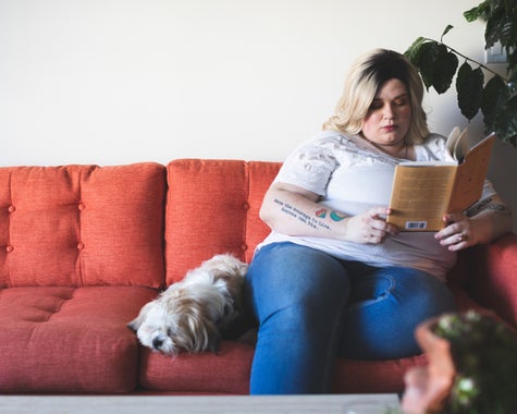 A woman sitting on a couch reading a book. Her small, fluffy, white dog is napping because it can't read, unfortunately.